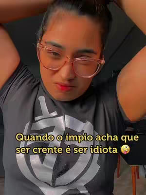 A woman with dark hair pulled back and wearing glasses is looking at the camera. She's wearing a black t-shirt with a white graphic. Text overlay in yellow reads: "Quando o ímpio acha que ser crente é ser idiota" with a sticking-out-tongue emoji.