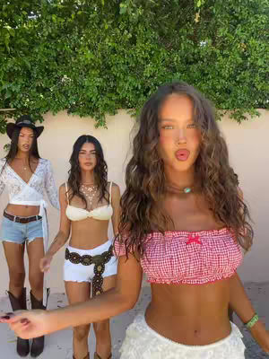 Three young women pose outdoors against a green hedge backdrop. The focus is on the woman closest to the camera, with long wavy brown hair, wearing a red and white gingham crop top and a white ruffled skirt. She's making a kissing face. Two other women stand behind her, slightly out of focus, wearing casual outfits. The lighting suggests a bright, sunny day.
