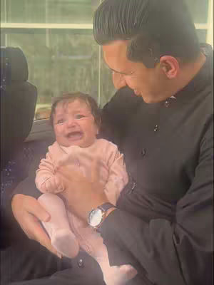 A man is holding a crying baby. The man is looking down at the baby with a gentle expression. The baby is wearing a pink bodysuit. The background is blurred but appears to be inside a vehicle with windows.
