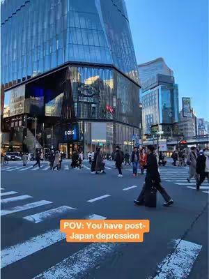 A street scene in Japan with a large modern building on the left, and a person walking towards the camera in the foreground. The caption text is in the bottom right corner.