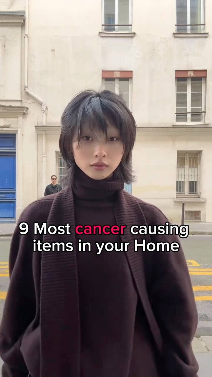 A woman stands in front of a multi-story building with windows. The woman has short, dark hair and is wearing a high-necked brown sweater. Text on the image states '9 Most cancer causing items in your Home'.