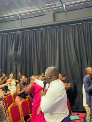 A live event in a church setting, with a man speaking into a microphone. The image features a crowd with hands raised in worship or praise. The setting is lit with overhead lights, and there are black curtains in the background. The scene has a strong emphasis on religious content, as indicated by the caption.