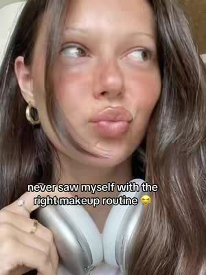 A selfie featuring a woman with fair skin, brown hair, and a soft, natural makeup look. She is looking to her side with a slight pout. She's wearing gold hoop earrings and her left hand is holding a ring. Headphones are around her neck. Text overlayed on the image reads, 'never saw myself with the right makeup routine'.