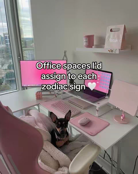 A well-lit, aesthetically pleasing home office setup. A dog is sitting in a corner. On the white desk, a laptop, keyboard, computer monitor, and desk lamp sit atop a white desk. The room is decorated in shades of pink and white. A window is visible in the background, with a city view.