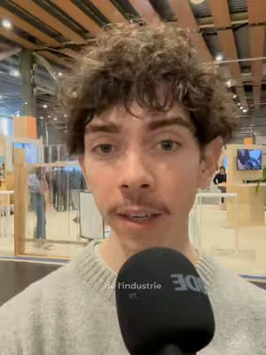 A young man with curly brown hair and a mustache is holding a microphone in front of his face. He is wearing a light gray sweater. The background is blurred but seems to be a conference or exhibition space.