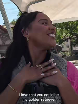 A selfie-style shot of a woman in a vehicle. She has long, dark hair and is smiling. Her arms are crossed in front of her chest, showing multiple rings and a delicate bracelet. There is text overlayed on the image.