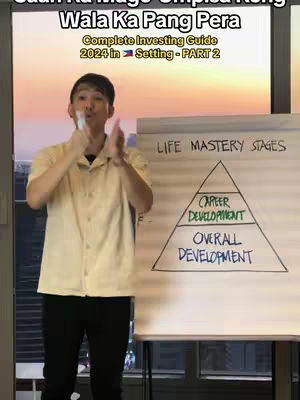 A man in a light yellow collared shirt and black pants stands in front of a presentation board with a whiteboard marker in his right hand, seemingly clapping. The presentation board has the words 'LIFE MASTERY STAGES' as well as a triangular diagram with the sections being 'CAREER DEVELOPMENT' and 'OVERALL DEVELOPMENT'. The background is blurred and shows the view through a window.