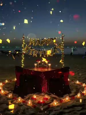 A romantic beach birthday setup at dusk. A table is set with a red tablecloth, surrounded by candles and rose petals. Above the table, gold balloons spell out 'HAPPY BIRTHDAY', illuminated by string lights. The background features the ocean, a dusky sky, and some fireworks.