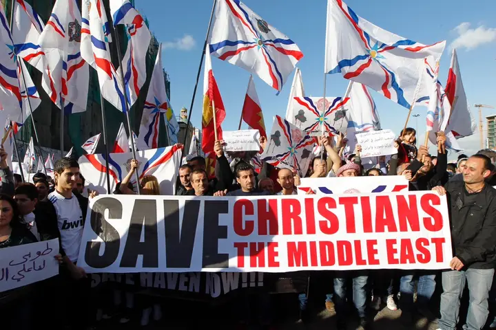 A large group of people march in a city street, holding flags and banners. The flags are white with a red and blue emblem on them. A large banner in the foreground reads "SAVE CHRISTIANS THE MIDDLE EAST". The sky is blue and the lighting appears to be daylight.