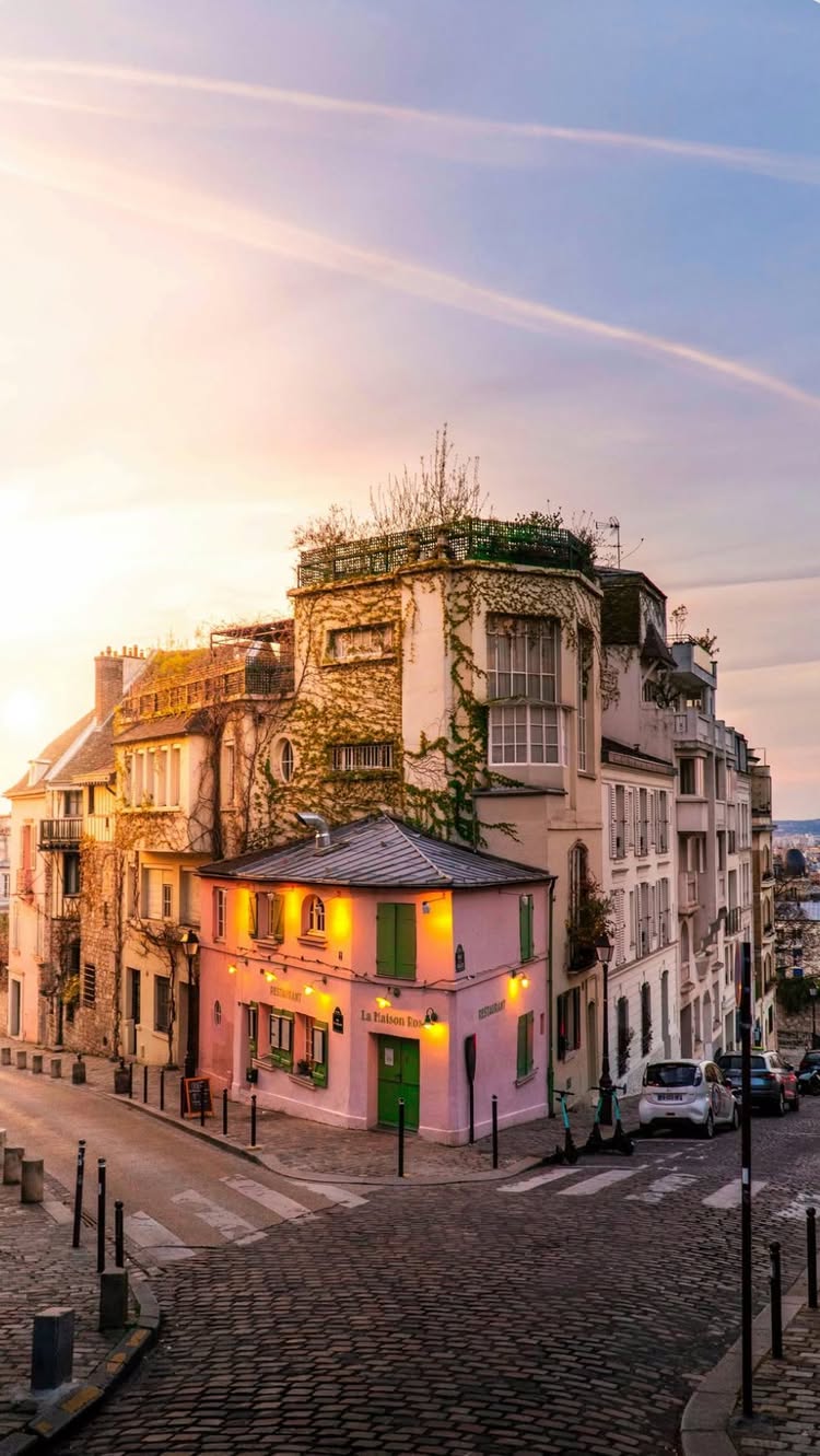 A picturesque street scene in Paris at sunset. Buildings of various heights and architectural styles line a cobbled street, with a pink restaurant at the corner. Soft sunlight bathes the scene, creating long shadows and highlighting the textures of the buildings and cobblestones. The sky transitions from blue to pastel colors, adding a warm glow.