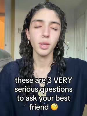 A selfie-style video showing a young woman with long, dark curly hair and a nose ring. She is looking down with her eyes closed, suggesting a reflective or emotional moment. Text overlayed on the image reads 'these are 3 VERY serious questions to ask your best friend' followed by an emoji. The background is soft, with neutral tones.