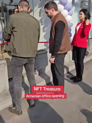 The image captures an office opening event. Three people are shown, two men and a woman. The main subjects are in focus, standing just outside a glass door, with a red ribbon stretched across. Behind the individuals, a decorative white and purple balloon arrangement is visible and sunlight casts shadows. The men are dressed casually, and the woman stands with a red jacket. Below, a red banner states "NFT Treasure" and "Armenian office opening".