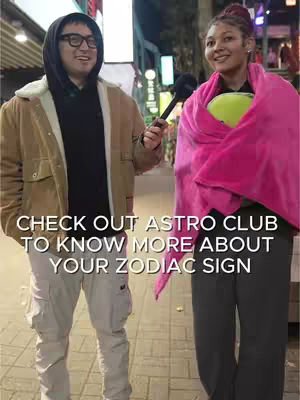 Two people are standing on a street, apparently conducting an interview. A man on the left, wearing glasses and a hooded jacket, holds a microphone. A woman on the right, wrapped in a pink blanket, is holding a yellow object. The background shows city lights.