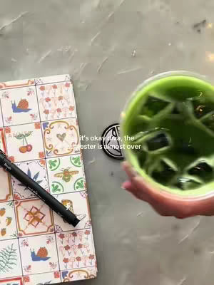 The image shows a top-down view of a table. On the left side, there is a patterned notebook lying open, next to a black pen. To the right, a hand is holding a clear glass filled with green liquid and ice cubes. The top of the glass is visible. In the center, a circular sticker with a design is placed on the table. The background appears to be a marble or stone surface.