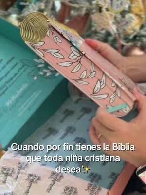 A person’s hands holding a pink Bible with white leaf pattern on a spiral-bound cover, with a teal and white box in the background. Overlayed text reads in Spanish.