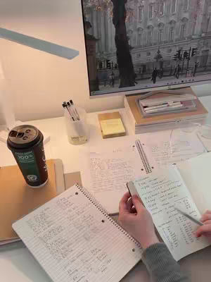 The image shows a study desk with various items on it. The desk is mostly white with natural lighting. On the desk, there's a coffee cup, notebooks, pens, and a monitor in the background displaying an image. A person's hands are visible, holding a notebook and writing.