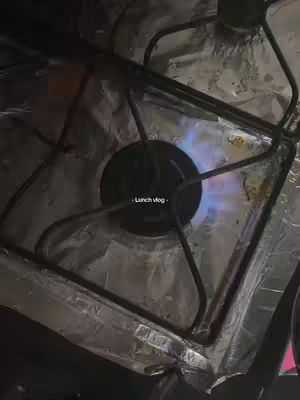 An overhead shot of a gas stove burner with a blue flame. The burner is surrounded by a metal grate and covered with aluminum foil. The text "Lunch vlog" is placed near the center of the black burner.