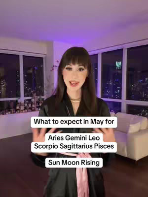 A woman with long dark hair and a black outfit stands in a well-lit room. Text overlays appear on white rectangles, introducing topics about astrology, including what to expect in May for certain zodiac signs. Background includes windows overlooking city lights and basic furniture.