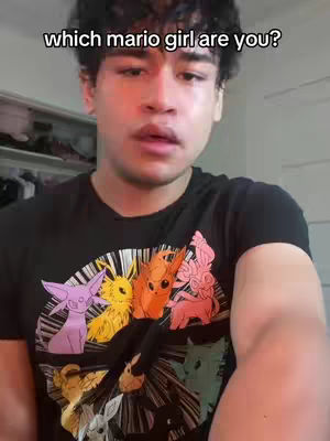 A selfie-style video cover featuring a young man looking directly at the camera. The man wears a black t-shirt with a colorful graphic of several cartoon creatures. Above the man's head is overlaid text in a bold white font, reading "which mario girl are you?". The background is a softly lit, plain interior, suggesting a casual setting.