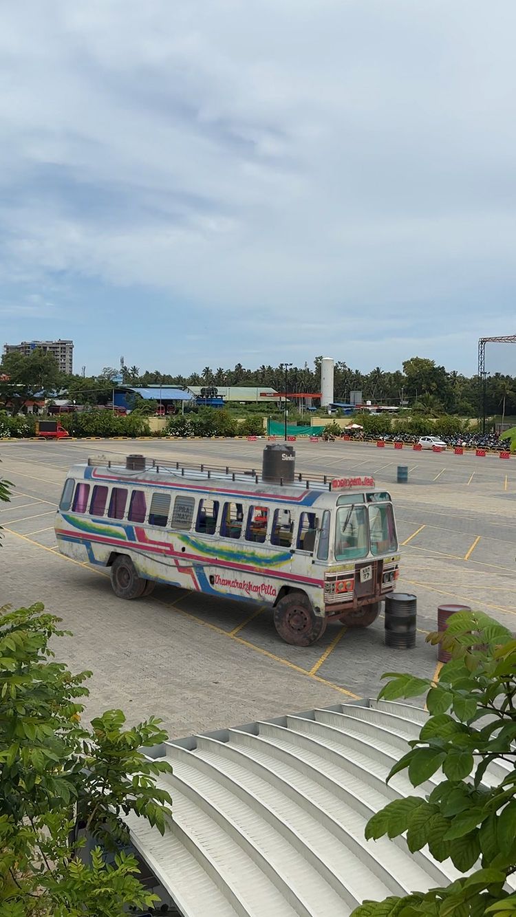An eye-level shot of a retro bus parked in a large paved lot. The bus is off-white with colorful stripes and lettering that reads  "Thamarakshan Villa." The bus has tinted windows and a water tank on top. Surrounding it are empty parking spots, a few barrels, and some vehicles parked in the distance. Lush green foliage and part of a building frame the foreground. The sky is overcast.