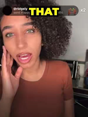 A woman with green eyes and curly dark hair is looking at the camera with a surprised or excited expression and her mouth open. She is touching her cheek with her left hand. The background is slightly blurred, suggesting an indoor setting.