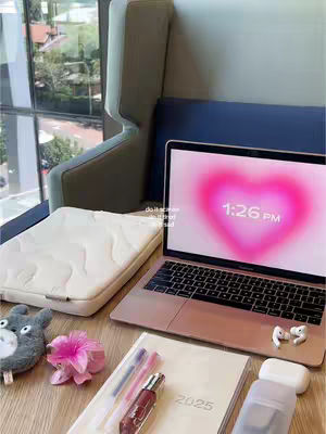 A well-lit, top-down view of a desk setup. The primary focus is on a rose-gold laptop with a bright pink heart-shaped image on the screen. The desk is covered in items suggesting study or work: a cream-colored laptop sleeve, a notebook, pens, a small perfume bottle, a small stuffed animal, a pink flower, and wireless earbuds. The background includes a window with a blurry view of greenery and a sleek, curved chair. The overall vibe is clean, modern, and aesthetic, with a focus on soft colors.