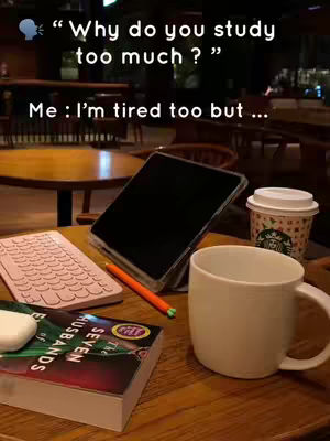 A close-up shot of a wooden table in what appears to be a cafe or study area, with various items scattered on it, including a tablet with a dark screen, a keyboard, an open book titled 'The Seven Husbands', a coffee cup, a white mug, and a Starbucks cup. The lighting suggests an indoor setting, possibly at night due to the dim lighting. Text overlays are added to the image.