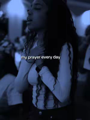 The image is a medium shot featuring a young woman with long, curly dark hair. She is wearing a white long-sleeved shirt and is positioned with her hands clasped over her chest. Her eyes are closed as if she is in prayer. The background is blurred, suggesting a gathering or a public space due to the presence of other blurred figures. The color palette leans towards cool tones, with a dominant blue hue.