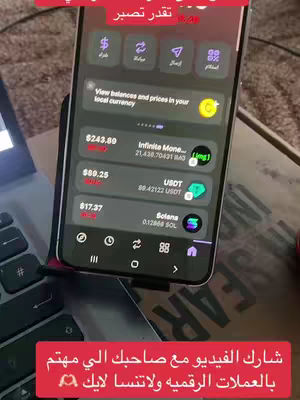 A phone displays a cryptocurrency app interface, held vertically against a dark setting with a laptop keyboard in the foreground. The phone screen shows various cryptocurrency values displayed on the screen. Two text overlays are visible, one at the top and the other at the bottom of the image.