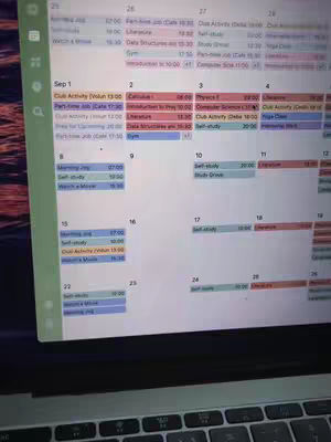 A laptop screen displaying a calendar application interface. The calendar is organized with dates and scheduled events, shown in a mix of colors such as blue, red, and orange for event markers. The screen is positioned at an angle, with a mostly visible keyboard in the foreground, suggesting the user is working with the application. The background is blurred, putting emphasis on the calendar view. 
