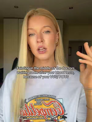 A young woman with blonde hair is speaking directly to the camera. She is wearing a white t-shirt with a colorful graphic. Text overlayed on the image reads: Fainting in the middle of the doctors waiting room after your blood tests, because of your VVS/POTS