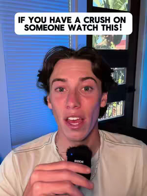 A young man with dark hair and fair skin looks directly at the camera against a blurred background. He holds a microphone with both hands in his lap as he speaks. Above his head is a transparent text box with white font. Additional background elements include vertical blinds on a blue tinted window.