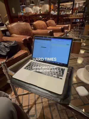 A laptop on a small metal and glass side table in front of a blurred background. The laptop is open, displaying a blue screen with highlighted text. Overlaid on the screen are the words "HARD TIMES", and below this, the words "gonna take it down and laugh when u cry". The background is a dimly lit room, with what appears to be a bar in the background, leather armchairs are visible too.