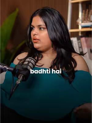 A woman with long black hair sits in front of a microphone, looking slightly off-camera. She's wearing an off-the-shoulder teal top. The background is somewhat blurred, with hints of bookshelves and plants. The image has a natural lighting setup.
