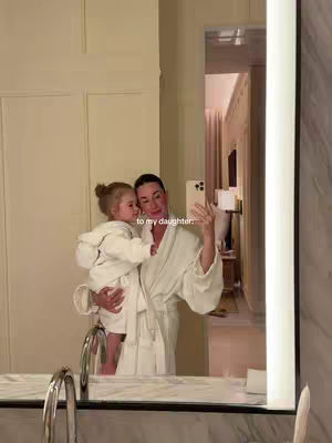 A woman holds a young child in her arms in front of a mirror. Both are wearing white robes. The woman is taking a selfie with her phone. The background shows a well-lit bathroom.