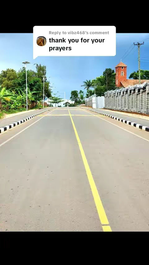 A wide shot of a road extending into the distance. A yellow line runs down the center, with trees, a church, and a blue sky in the background.