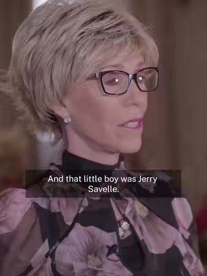 A woman with short, light-colored hair and glasses is speaking directly to the camera. The background is slightly blurred, suggesting an indoor setting.