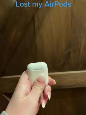 A hand holding a white AirPods case, with the text "Lost my AirPods" in blue at the top. The background is a brown, textured wooden surface.