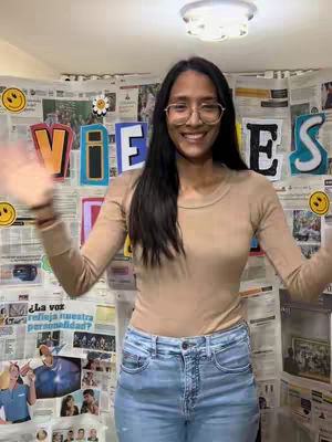 A woman wearing glasses and a light brown long-sleeved shirt and jeans stands in front of a wall covered in newspaper clippings and colorful letters. She is smiling with her arms out to the sides, palms up.