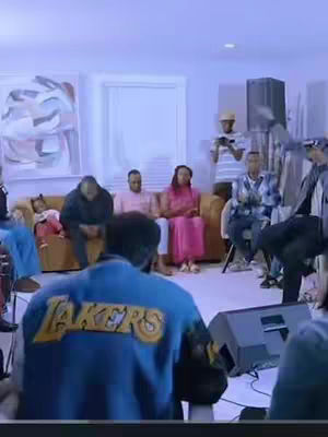 The image captures people in a room possibly holding a worship session. Foreground shows a person wearing a blue Los Angeles Lakers jacket. There are several people in the background sitting on a couch.