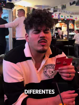 A man with curly hair holds two red cards in the foreground with a bar in the background. The man is making a face. In the background, other people are visible, and various flags are hanging.
