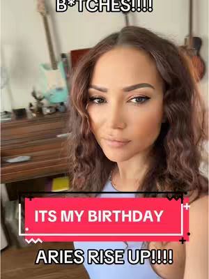 A selfie-style shot featuring a woman with long, wavy brown hair. She's looking directly into the camera with a slight smile. A bold, red rectangular overlay at the bottom says "ITS MY BIRTHDAY" and "ARIES RISE UP!!!!". The background is blurred, showing interior details including a guitar.