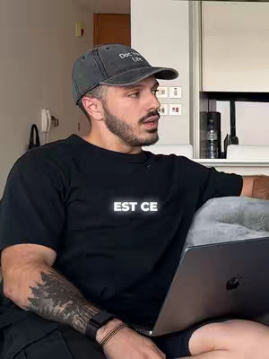 A man in a black t-shirt and a baseball cap is sitting on a couch with his laptop. The shirt has the text "EST CE" on it. He is looking to the side, possibly speaking or listening. He has a beard and a tattoo on his arm.