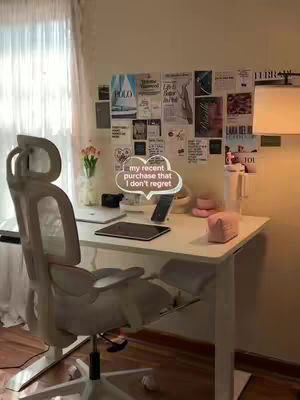 A well-lit, neutral-toned workspace featuring a white desk, office chair, and various items. Behind the desk, a wall is adorned with a collection of papers and prints. A large window with sheer curtains is visible on the left. In the foreground, a modern white office chair is positioned, suggesting a focus on comfort and functionality.
