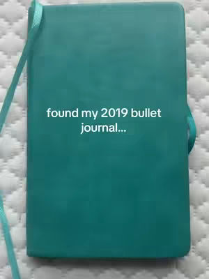 A teal bullet journal lies open on a white quilted surface. The journal has a matching teal ribbon bookmark. The cover image has white text overlaid.