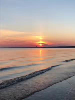 A scenic view of a beach at sunset or sunrise. The sky is a gradient of colors from blue to orange and yellow. The sun is near the horizon, partially obscured by clouds. The water is calm, reflecting the colors of the sky. The waves near the beach are small.