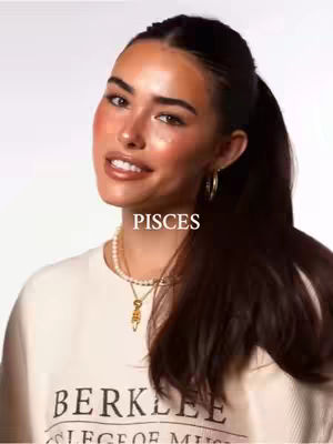 Close-up shot of a woman with long dark hair pulled back in a high ponytail, wearing a cream-colored sweatshirt. She is smiling with a hint of a blush, wearing gold hoop earrings and a pearl necklace, with text overlayed that says \"PISCES\".