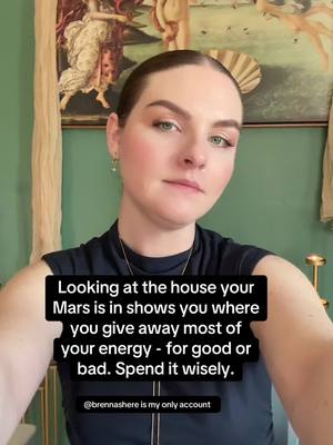A woman with light skin and dark hair is centered in the frame. She's wearing a black top with a gold necklace. The background is a wall painted in a light green color, with a painting and draping fabric on the wall behind the woman. The text says, "Looking at the house your Mars is in shows you where you give away most of your energy - for good or bad. Spend it wisely." with the username @brennashere is my only account.