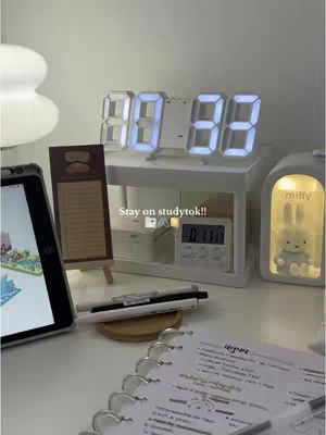 A well-lit desk setup with a digital clock displaying 08:33, an iPad showing a colorful image, a wooden easel holding a note, pens, a notepad with writing, and a light-up Miffy figurine. The overall aesthetic is clean and minimalist.
