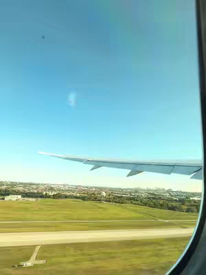 View from an airplane window. The wing of the plane is visible, stretching across the frame from the right towards the left. Below the wing, there is a landscape with green fields, a runway, and buildings in the distance. The sky is a bright blue, and the window frame is on the right edge of the frame.